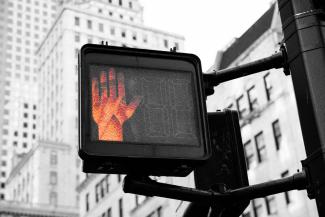 A crosswalk sign in a city. A red hand on the sign is lit up, signaling people to stop