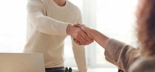 Man and woman shaking hands over a desk. On the desk is a laptop and a coffee cup.